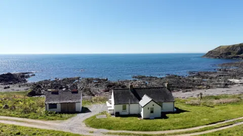 Galbraith A white cottage and hut next to it on the shoreline looking out to sea. A track runs behind the two buildings