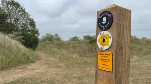 At the forefront of the photo on the right is a post, with three signs on. The top is a black circle, reading 'King Charles III England Coast Path.' The sign underneath is a white circle with a yellow arrow which is pointing to the left, also reading 'King Charles III England Coast Path.' The bottom line reads 'Public Right of Way.' In the background, to the left, is a path surrounded by grass and trees. 
