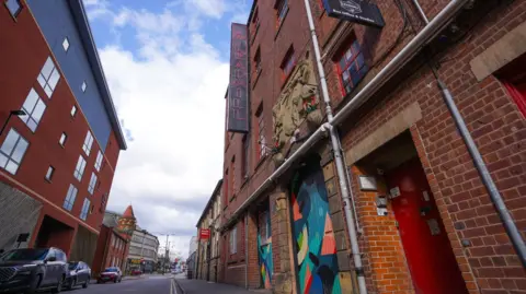 A street with a four-storey building  on one side. There is a black sign outside with the words 'The Leadmill' in red, neon writing. Two doors to the building are painted in multi-colours. 