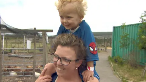 A woman is walking with her young son on her shoulders. She is smiling and wearing glasses and has light brown hair. Her son is blonde and wearing a blue and red Spiderman t shirt. There are chickens in a field to their right