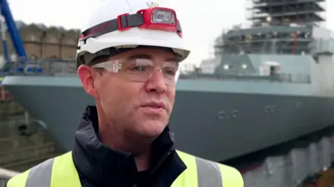 BBC A man in a florescent jacket wearing a white hard hat with goggles is standing in front of a large grey warship.