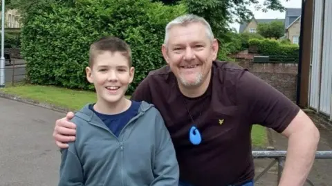 There is a young boy wearing a grey coat and blue pants. He has brown hair. He is being hugged by an older man with grey hair. He is wearing a maroon t-shirt, around his neck is a blue silicone toy. They are stood in front of some trees. They are both smiling. 