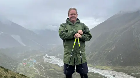 Simon Clark is standing on a mountain with the valley beind him. He is wearing a green jacket and leaning on his walking sticks.