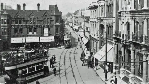 Ipswich Maritime Trust A black and white image of Ipswich Cornhill showing trams, tram marks, people walking about, a number of buildings all around the trams and awnings. 