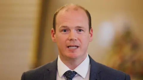 PA Media Gordon Lyons, a man with short fair hair, wearing a dark jacket, white shirt and blue tie with yellow pattern.