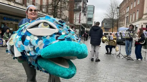 A woman is wearing a large padded fish costume in Bedford Square in Exeter with brass band players in the background.