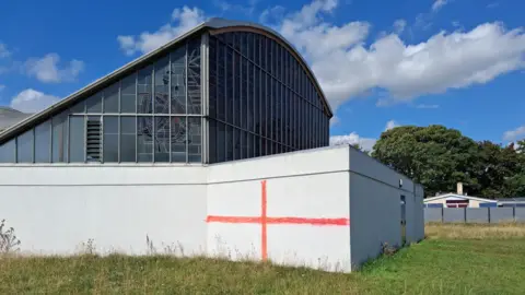 LDRS There is a white building with windows above. There is a red cross painted on a white wall. There is also long grass and trees in the distance.