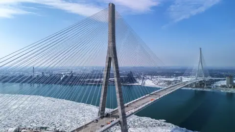 The Gordie Howe International Bridge, a cable-stayed bridge featuring six traffic lanes, is seen in a drone image. The ground on either side is covered in snow, with unfrozen water flowing beneath. 