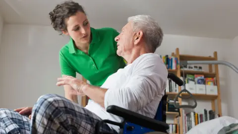 A woman in a green t-shirt is talking to a man in a wheelchair. She has her brown curly hair pulled back in a ponytail. He has grey hair and is wearing checked trousers and a long-sleeved white top. They are probably pyjamas.
