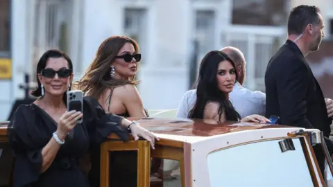 Reuters Kris Jenner, in dark dress and sunglasses, with sparkling bracelet and earrings, smiles while stood at the stern of a small motorboat. Khloe and Kim Kardashian, appearing similarly glamorous are also on board, as are two unknown men, one of whom wears a security guard's earpiece.