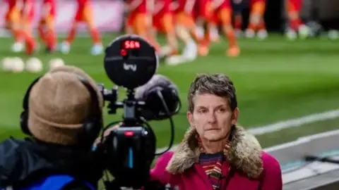 FAW Laura McAllister stands in front of a cameraman holding a large camera on his right shoulder. A football pitch can be seen in the background behind her. She wears a pink coat with a fur trim collar and has short brown hair.