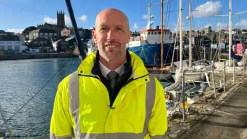 Stuart Reid is standing n the quay on Penzance. He is weering a high-viz jacket over a shirt and tie. He has a blad head and beard and is smiling. Behind him a church is across the water and there are boats tethered to the quay.