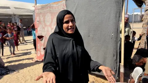 BBC Sylvia al-Shurafi, a woman wearing all black, pictured in front of a make-shift divider constructed from beams and two large pieces of fabric with some tears in them. She has a serious expression and has her hands out in front of her. Behind her a group of people including children looks on.