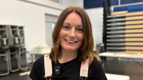 Ben Schofield/BBC Laura Deller smiling and looking directly down the camera. She is wearing a black top and the shoulder straps and buckles of a pair of pink dungarees can also be seen. She has straight, shoulder-length hair. Laura is standing inside a large room, with stacked chairs behind her on the left and a table to the right.