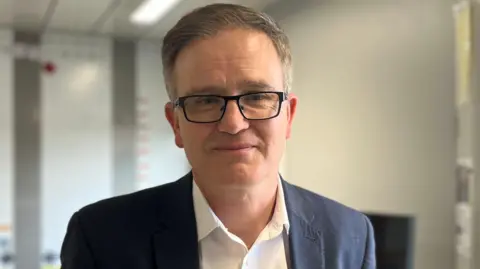 Sam Burton/BBC Chris Griffin, a man who is wearing glasses and is looking at the camera and smiling. He is wearing a white shirt and black suit jacket.