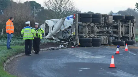 A cement mixer on its side at a roundabout. Two traffic police officers and a recovery driver, all in hi vis, stand beside the crashed vehicle.