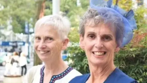 Supplied Wendy and Christina Read now. Wendy has short white hair and is wearing a white top with a purple and blue trim. Christina has curly grey hair and is wearing a fancy blue hairpiece made from lace and feathers and a blue coat