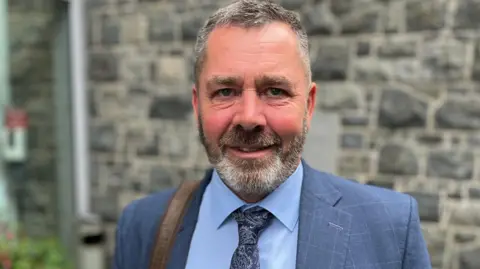 Deputy Marc Leadbeater - A man with black and grey hair and a a similar beard. He is wearing a blue suit with a blue shirt and a blue tie. 