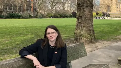 Transport for All chief executive Caroline Stickland sat on a park bench with Westminster in the background.