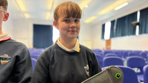 A girl in a navy school sweater with rows of blue chairs behind her, holding a pouch with the name Arwen scribbled on it