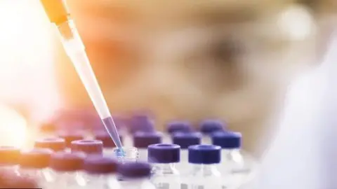 A pipette is pictured lifting liquid from a test tube. The tube is surrounded by many other tubes with purple lids.