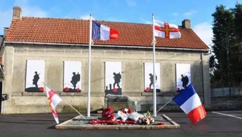 A rectangular building is seen with a tiled roof, on which silouhettes of soldiers are pictured with backpacks, poppies at their feet, and in front a memorial to those who lost their lives, as well as the flags of Guernsey and France. 