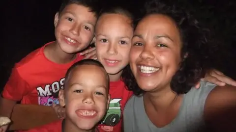Kerry Watson A close-up picture of Kerry Watson with her three sons, all of whom are smiling at the camera. Picture was taken in the early 2010s when children were junior school age. She is wearing a grey vest top and they are wearing red t-shirts.