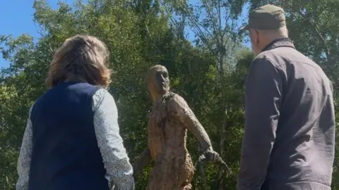 Jamie Niblock/BBC A man and a woman with their backs turned away from the camera look at a bronze statue of a man in a field on a sunny day.