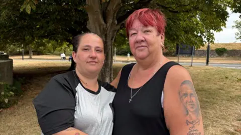 Shannon and Bev stand looking at the camera, with their arms around each other. Shannon wears a grey and white t-shirt with a black trim, while Bev wears a black vest. A tattoo of a face can be seen on Bev's left arm. There is a tree, grass and a road in the background.