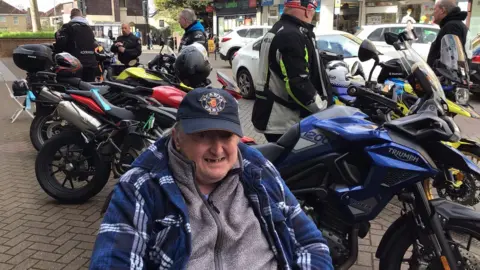 Westbury Court Care & Nursing Home A man in a fleece and blue shirt and dark blue cap smiles with motorbikes lined up behind him