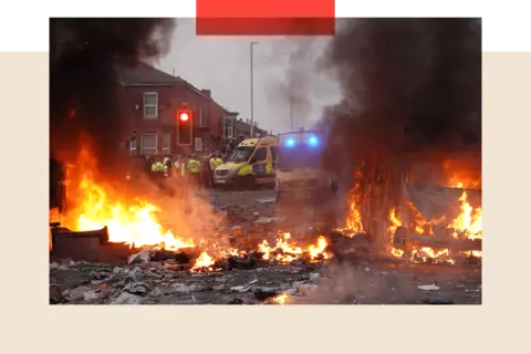 Getty Images Colour image of riot police holding back protesters. In the foreground a police vehicle is engulfed by fire.