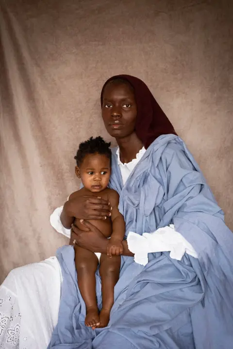 Dola Posh Dola Posh com um lenço preto na cabeça e um pano azul sobre um vestido branco olha fixamente para a câmera com uma expressão vazia. Ela está segurando a filha nas mãos.