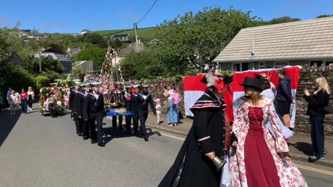 People are dressed up as they walk along the road in the procession. HMS Raleigh members are walking along in uniform, carrying the boat structure. A Saint George's Cross flag is draped on a stone wall. 