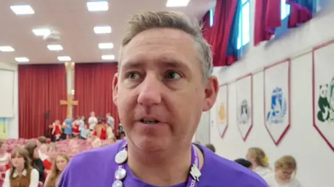 Year 6 lead Chris Cooper giving an interview while a lot of children are sitting behind him, waiting for show rehearsals.