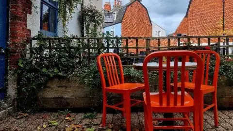 A striking, orange table and three chairs pictured to the right of the frame on a garden patio. there is a fence behind with trellis and ivy growing over it. Behind are houses of red brick