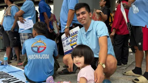 A girl in a pink shirt sits on the ground, in front of her father, wearing a blue polo shirt 