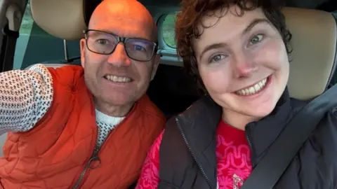 Lily McGarry A picture of Lily smiling while sat in a car. She is pictured next to her dad. He is bald and wearing black glasses.