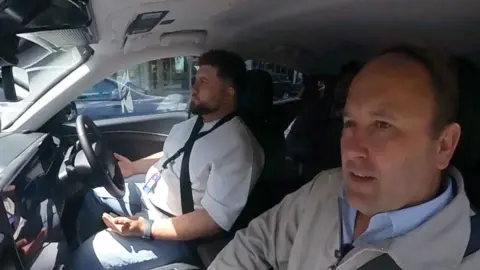 A safety driver is in the driver seat. His hands are off the wheel. A slightly concerned Transport Correspondent is in the passenger seat. 