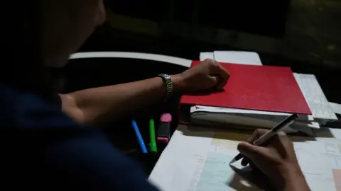 The Sachdev anachitis/BBC Anjali (name changed) is visible in a dark room that holds the pen, with documents scattered around it. 