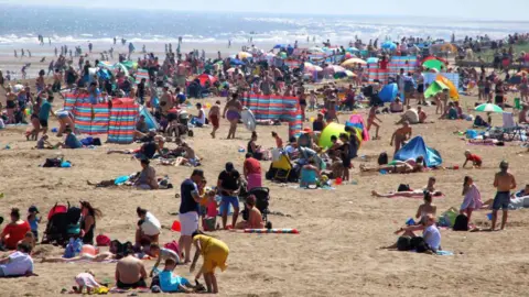 Families on Skegness beach, with crowds of people using windbreakers and beach umbrellas. The sea is in the background.