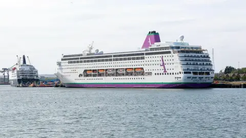 A large cruise ship, which is white, with purple and green markings, is seen docked at a port 