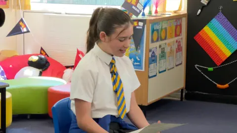 A mid shot of Valentina reading her letter from a piece of paper. She has tied back brown hair is wearing a school uniform, and is sitting on a blue chair. In the background are various toys and classroom decorations.