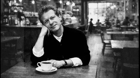 Paul Wolfgang Webster Wilson sits at a table with a teacup in front of him leaning his face on his palm. Shot by Webster