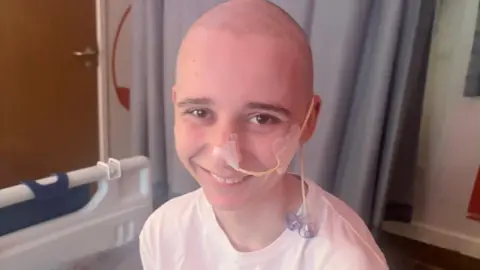 Family handout Leo sits on a hospital bed and has a hospital tube taped to his cheek and nose. He is bald and is wearing a white t shirt.