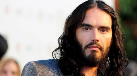 Reuters A head-and-shoulders photo of Russell Brand wearing a grey blazer and looking slightly past the camera.