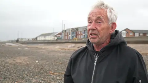 Philip Furnivall has white hair and wears a black zip up anorak