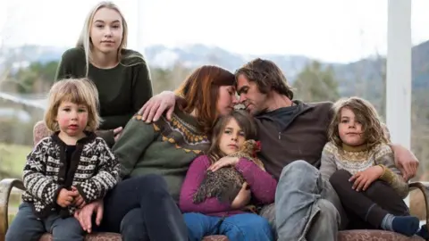 Maria and Nik Payne sit on a sofa leaning into each other and touching foreheads, as their four children sit around them