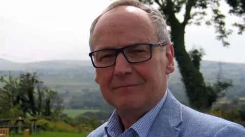 BBC Dyfrig Siencyn standing in a green field with hills and trees in the background. He has glasses on and is wearing a blue jacket and blue shirt