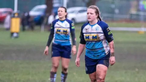 Newbury Rugby Football Club A woman wearing a purple and white rugby kit that is covered in mud. She's standing on a sports pitch and looking at something off camera. There's another player standing behind her in soft focus.