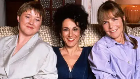 (L-R) Pauline Quirke, Lesley Joseph and Linda Robson sitting on a sofa smiling in Birds of a Feather